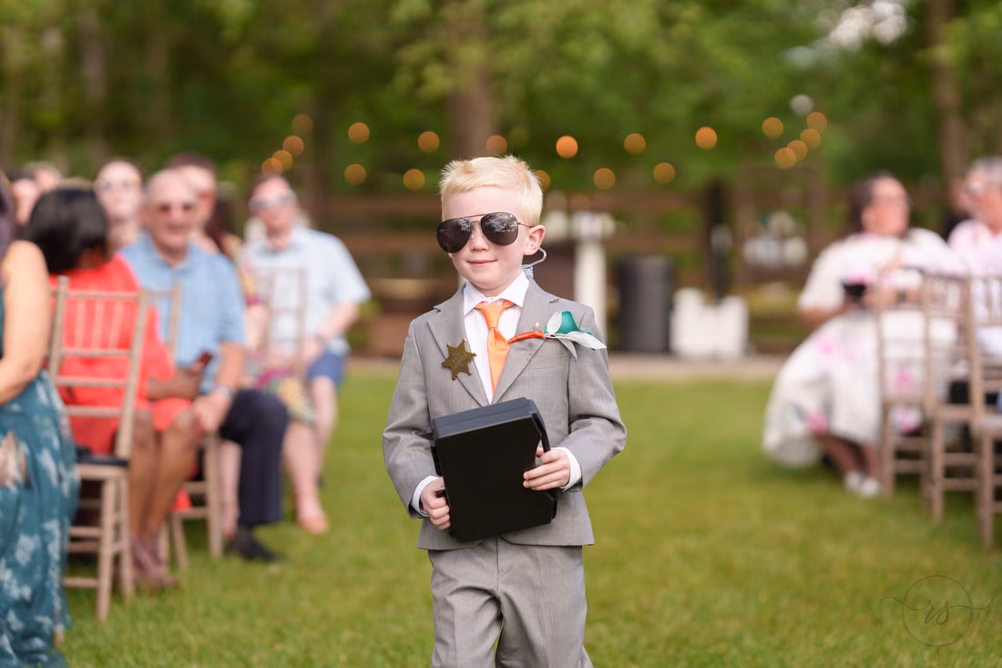 Ring bearer smiling in sunglasses as he carries the ring security case down the aisle at Wildhorse at Parker Farms
