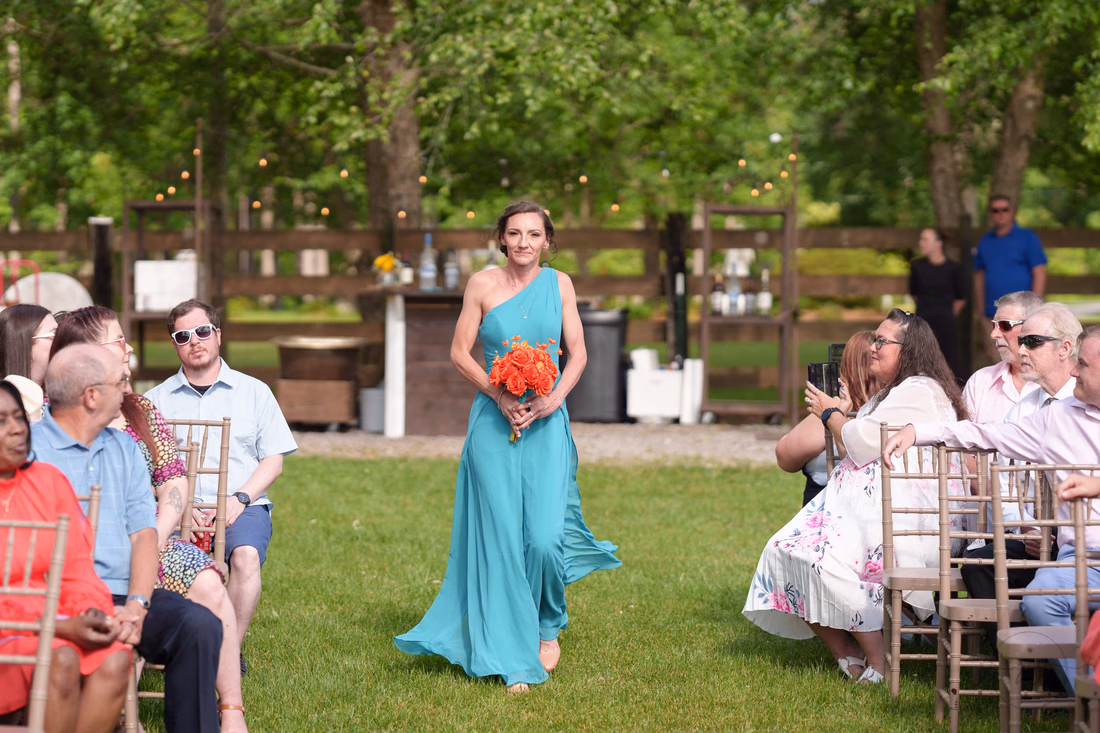 Bridesmaid in teal carrying orange roses down the grass aisle at Wildhorse at Parker Farms