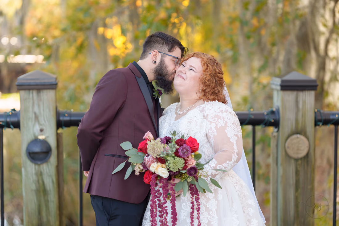 Groom kisses the bride's cheek by the railing in fall color on the Conway Riverwalk