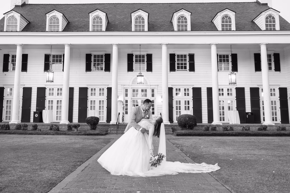 Black and white photo of the groom dipping the bride in front of the columned clubhouse at Pine Lakes Country Club
