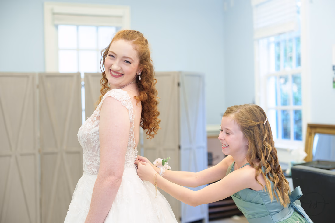 A flower girl fastens the back of the bride's dress at Pine Lakes Country Club