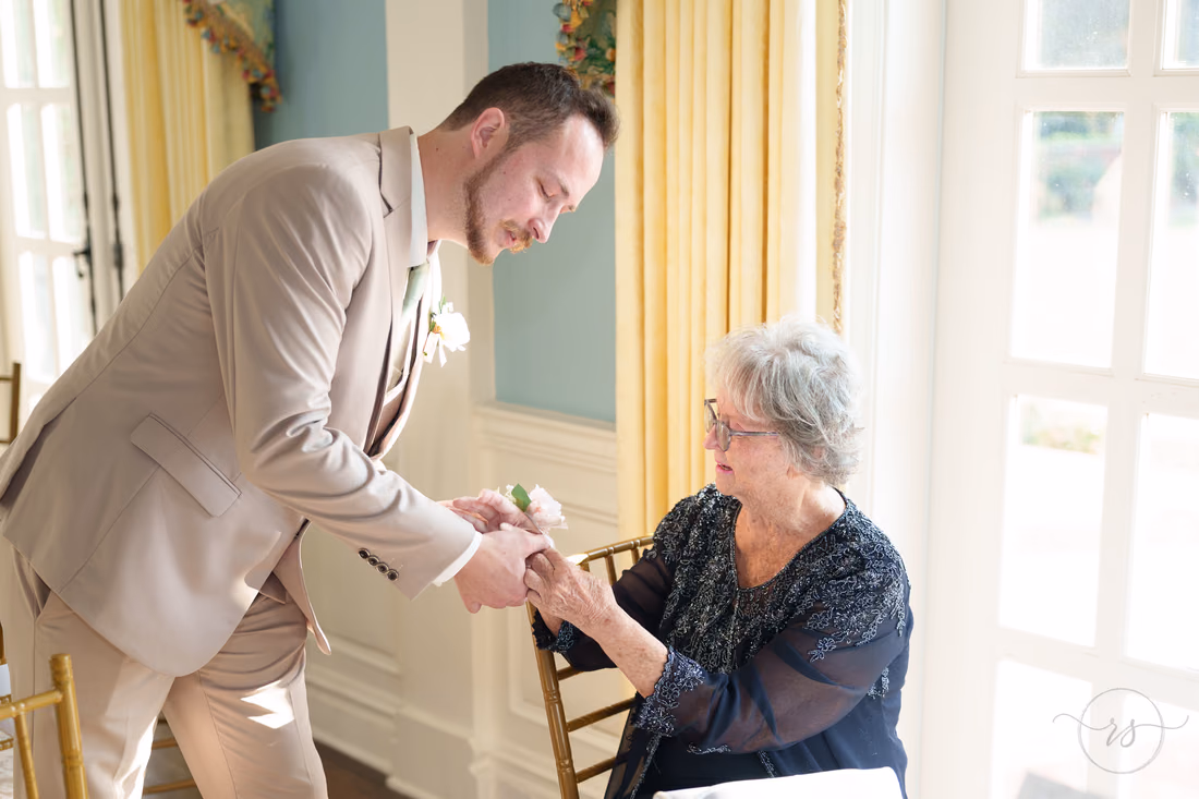 The groom leans down to an older woman seated with a flower in hand at Pine Lakes Country Club