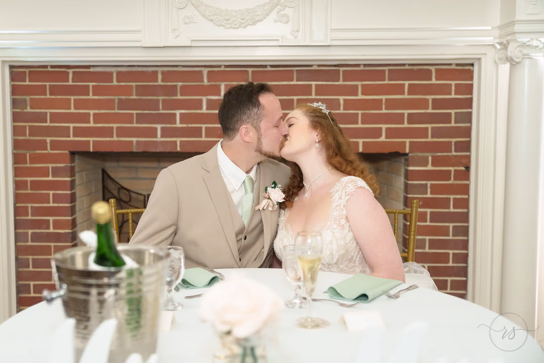 The bride and groom kiss at the sweetheart table by the fireplace at Pine Lakes Country Club