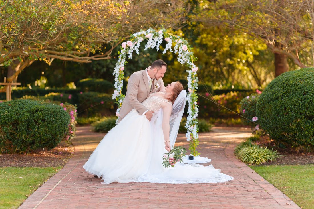 The groom dips the bride as they laugh under the flower arch at Pine Lakes Country Club