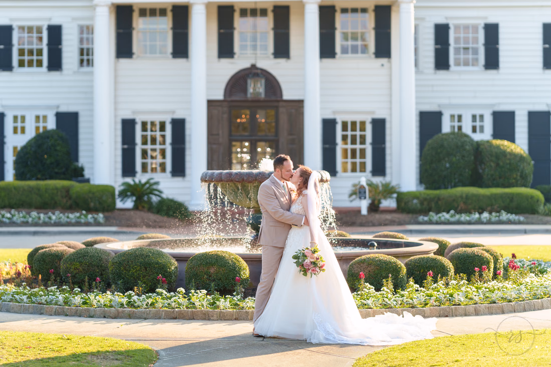 The bride and groom kiss by the fountain in front of the clubhouse at Pine Lakes Country Club