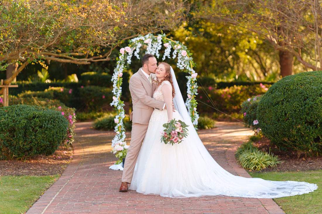 The groom kisses the bride's cheek under the flower arch at Pine Lakes Country Club