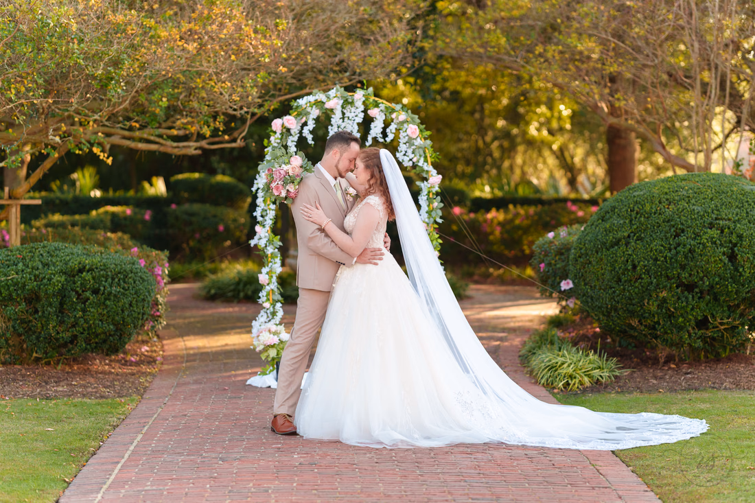 The bride and groom touch foreheads under the flower arch, her veil on the path, at Pine Lakes Country Club