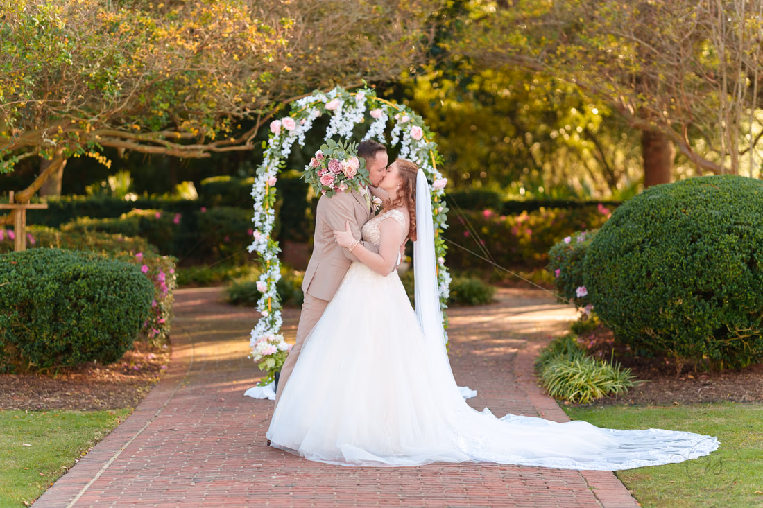 The bride and groom kiss, her bouquet behind his back, under the flower arch at Pine Lakes Country Club