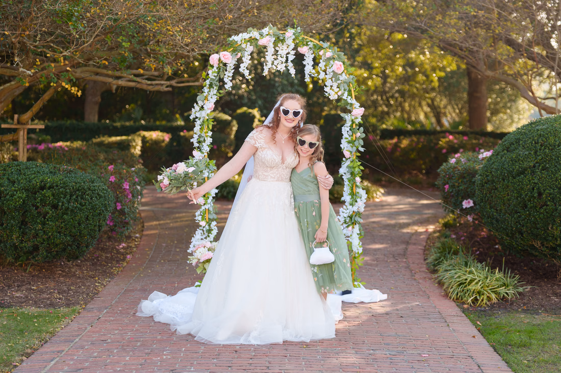 The bride and a bridesmaid in matching sunglasses under the flower arch at Pine Lakes Country Club