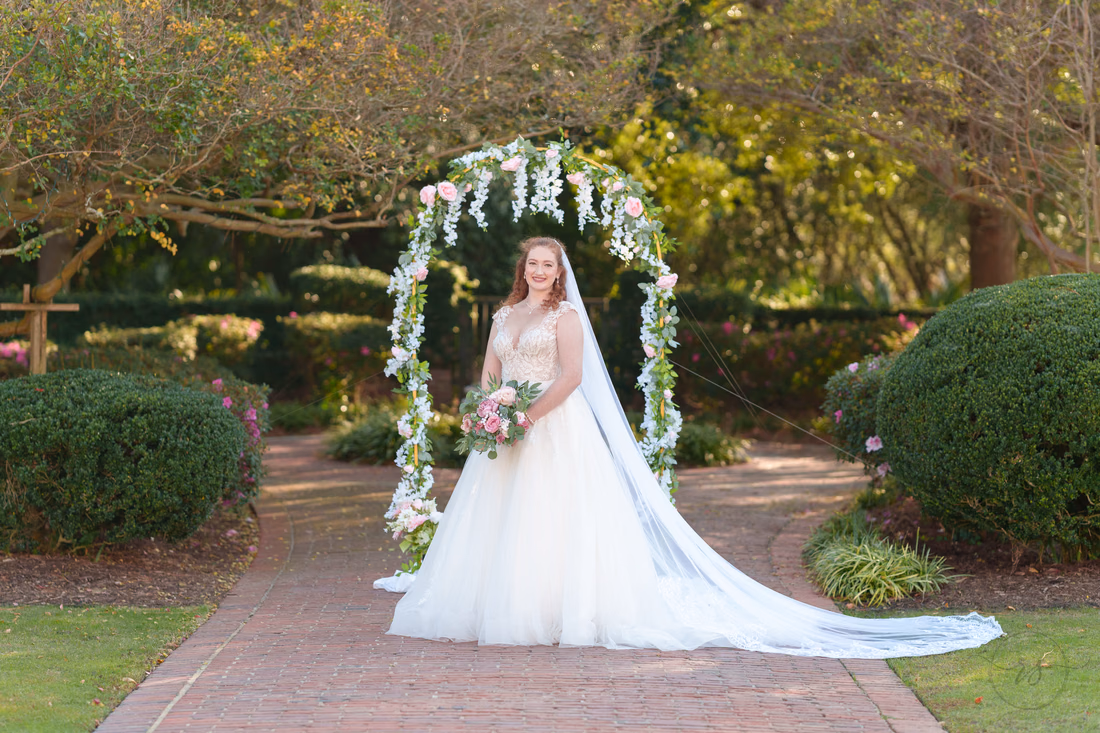 The bride's long veil spreads across the brick path under the flower arch at Pine Lakes Country Club