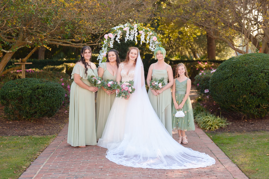 The bride with her bridesmaids and flower girl in sage green by the flower arch at Pine Lakes Country Club
