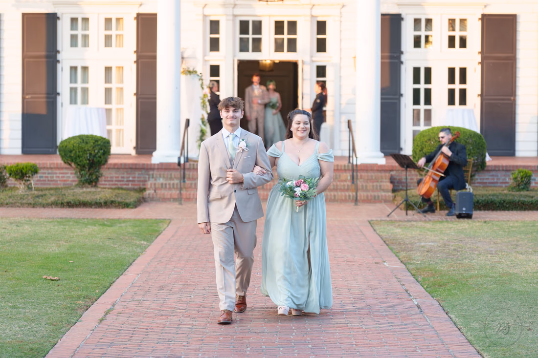 A groomsman and a bridesmaid walk down the aisle in front of the clubhouse at Pine Lakes Country Club