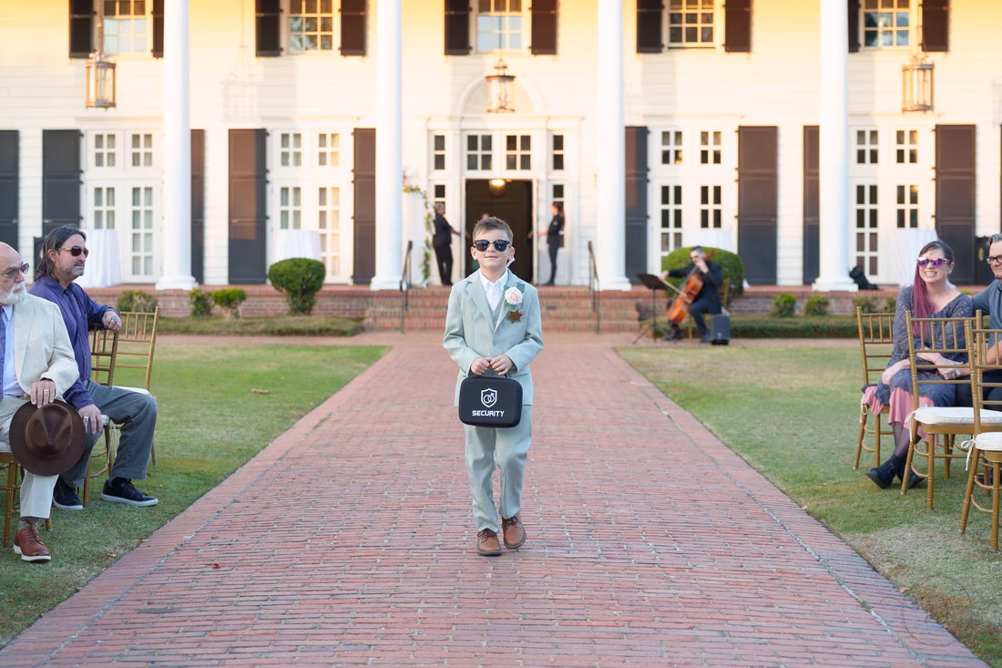 The ring bearer in sunglasses walks down the aisle carrying a small case at Pine Lakes Country Club