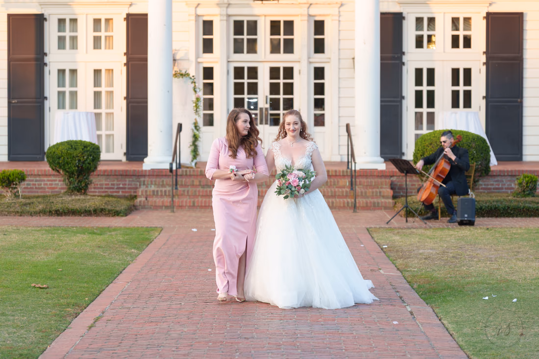 The bride walks down the brick aisle with a woman in pink at Pine Lakes Country Club