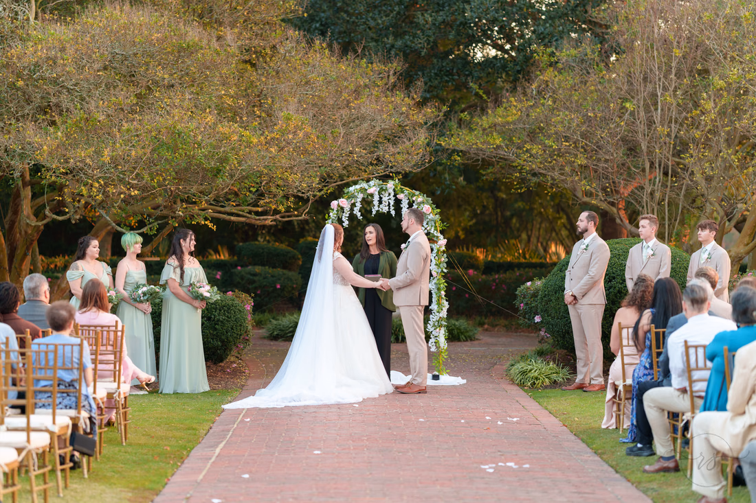 The bride and groom hold hands under the flower arch with the wedding party at Pine Lakes Country Club
