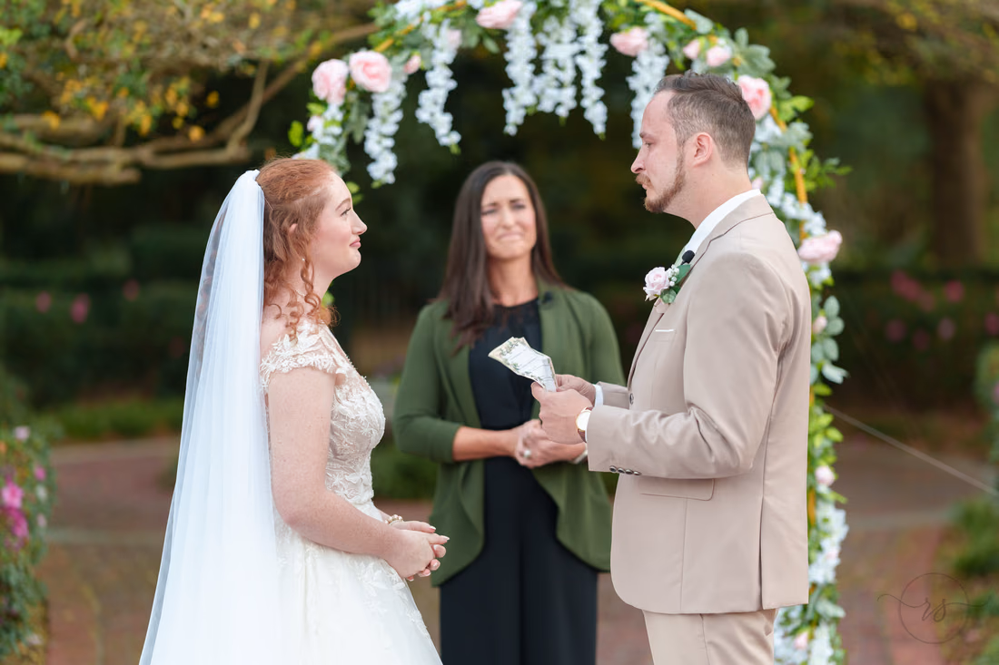 The officiant hands the groom the ring under the flower arch at Pine Lakes Country Club