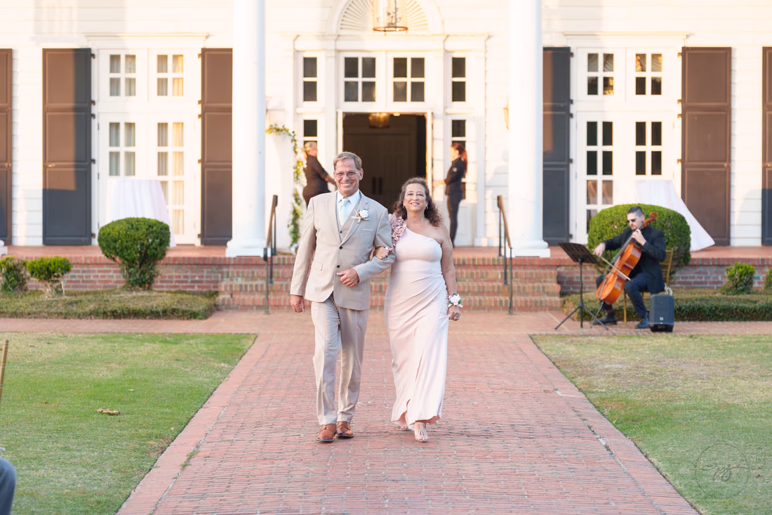 An older man and a woman in blush walk down the aisle as a cellist plays at Pine Lakes Country Club