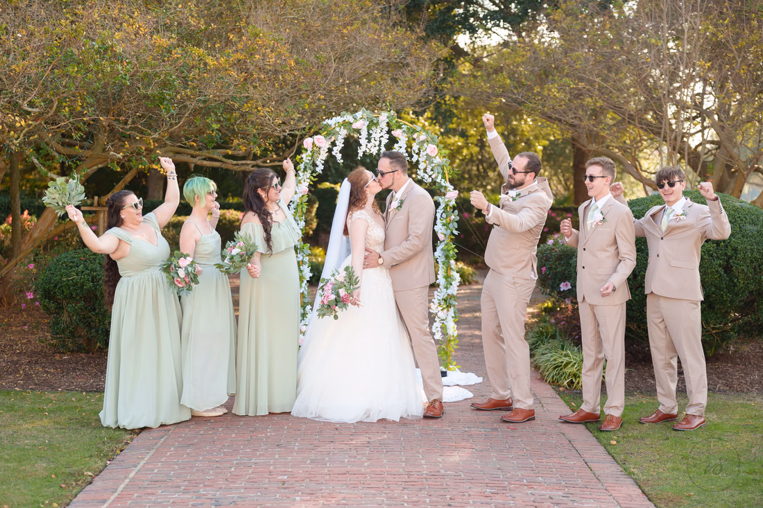 The wedding party cheers as the bride and groom kiss under the flower arch at Pine Lakes Country Club
