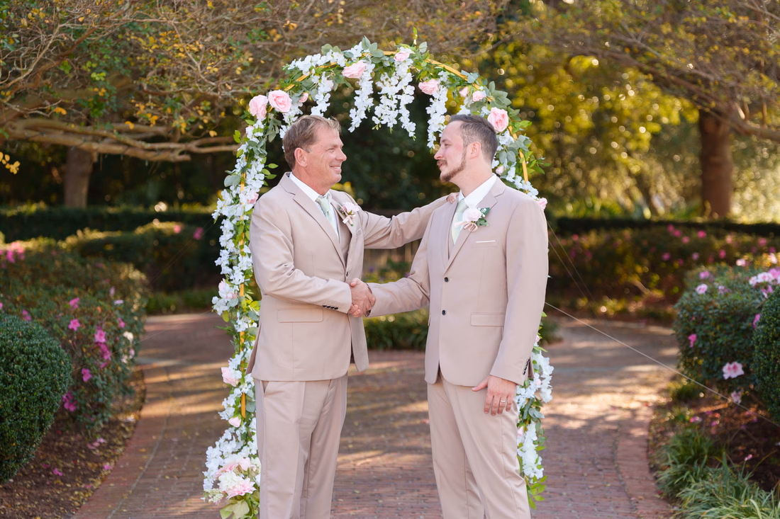 The groom shakes hands with an older man under the flower arch at Pine Lakes Country Club