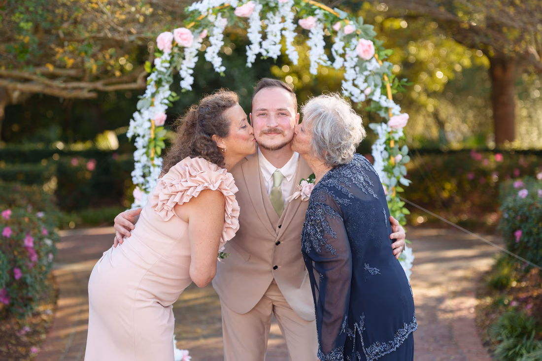 Two women kiss the groom on the cheeks under the flower arch at Pine Lakes Country Club