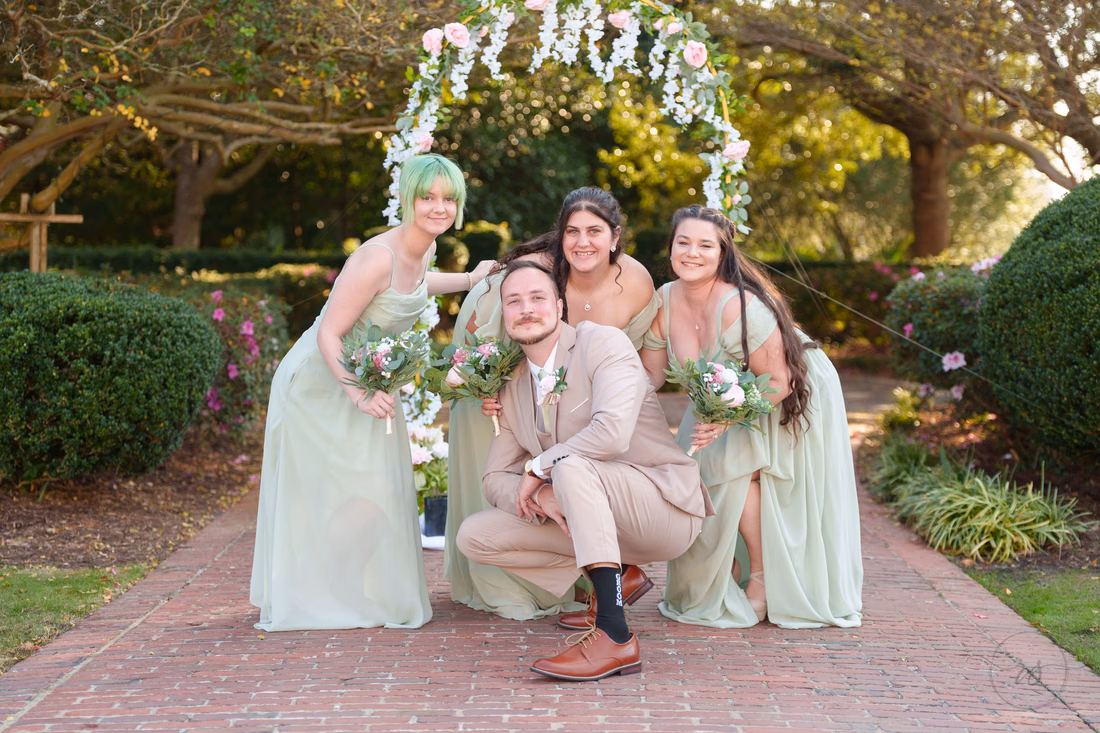 A groomsman crouches with the bridesmaids in front of the flower arch at Pine Lakes Country Club