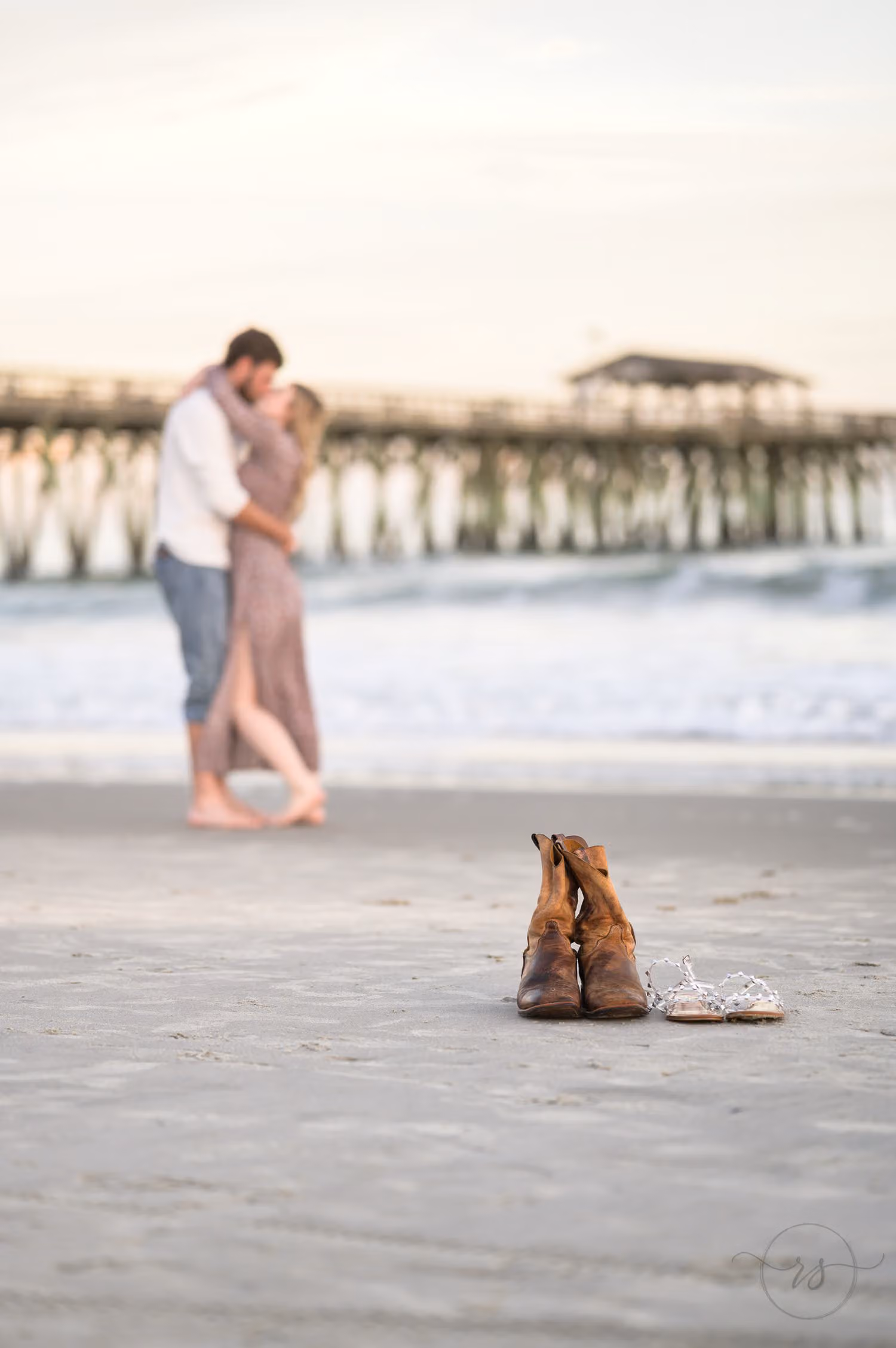 Pair of cowboy boots in focus on the sand while the couple embraces by the pier at Myrtle Beach State Park