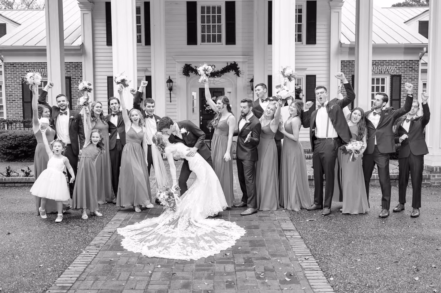 Bridal party cheering as the groom dips the bride by the columned clubhouse, in black and white, at Pawleys Plantation