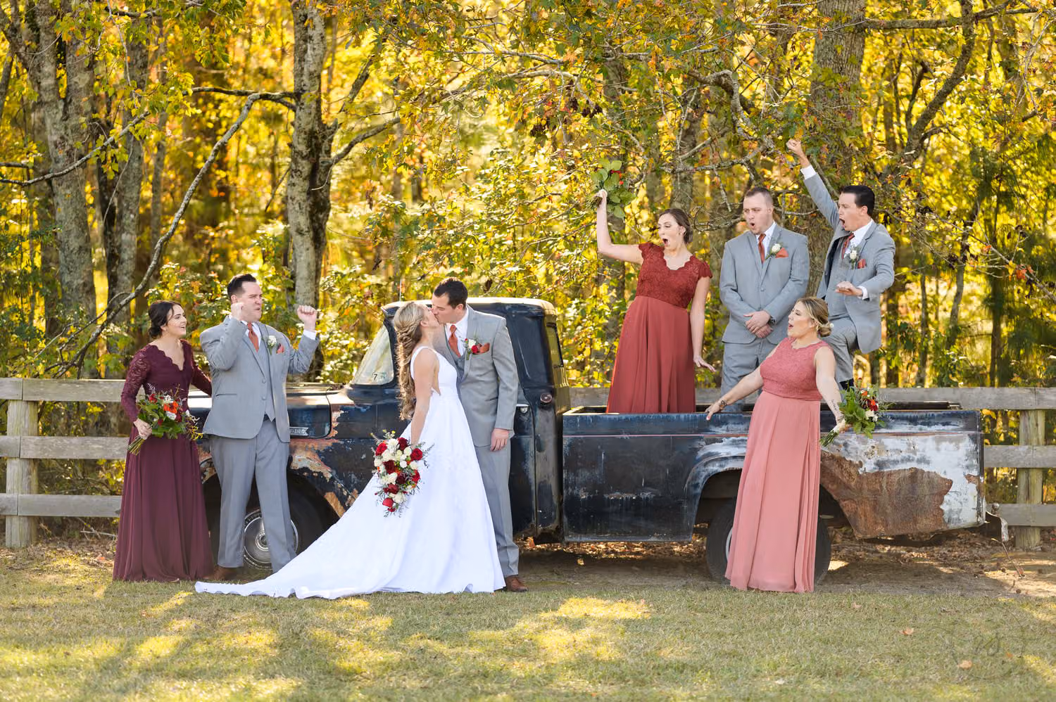 Bridesmaids in burgundy and rose and groomsmen in gray cheering around the couple by an old truck at The Blessed Barn