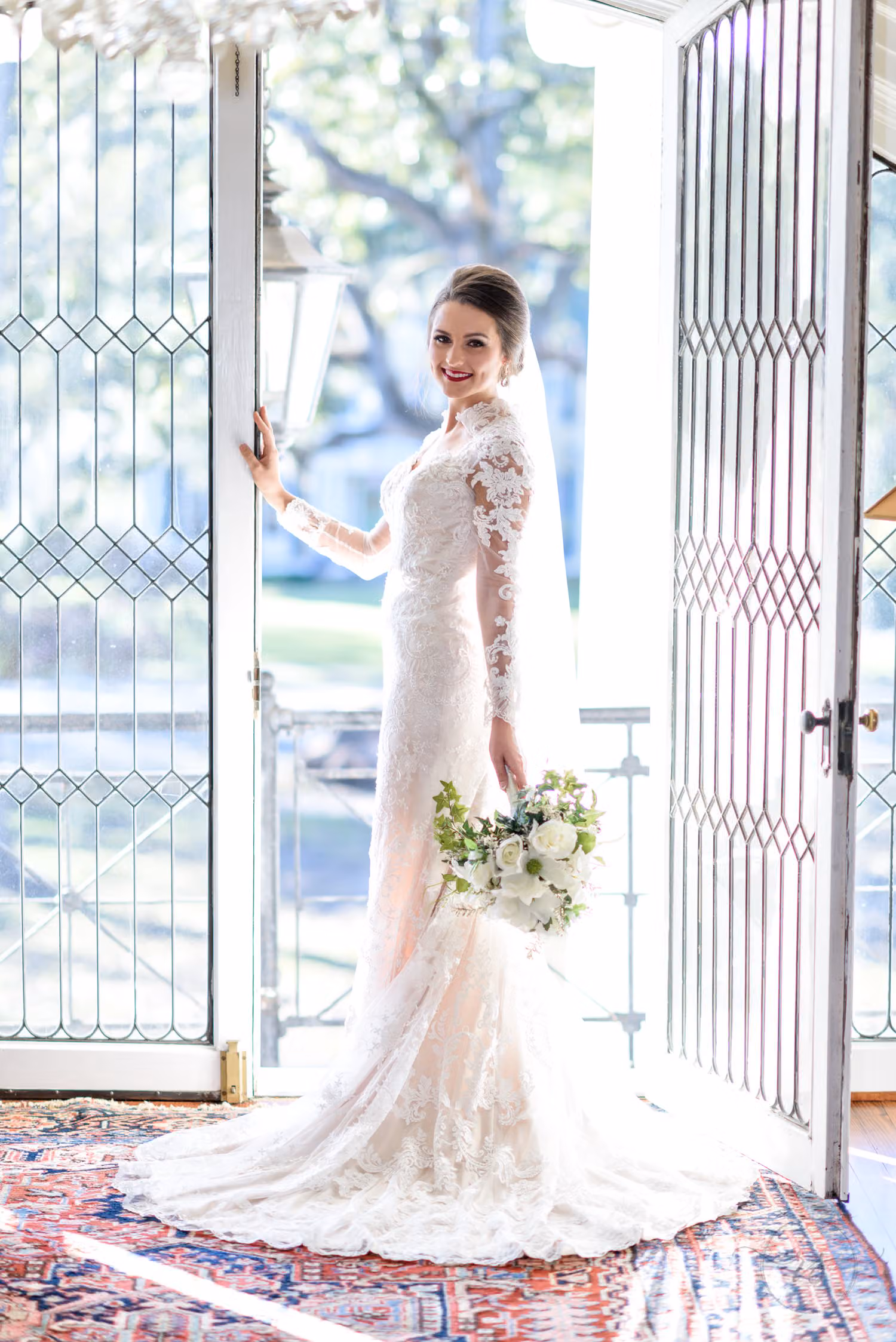 Bride in a lace gown with a white bouquet standing in an open glass doorway on a patterned rug at Rosewood Manor in Marion