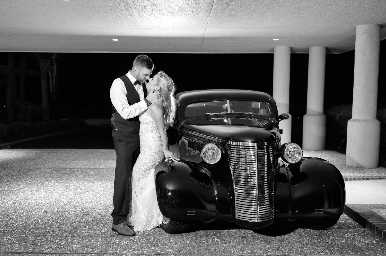 Black and white of the bride and groom kissing beside a vintage car at night at Dunes Golf and Beach Club