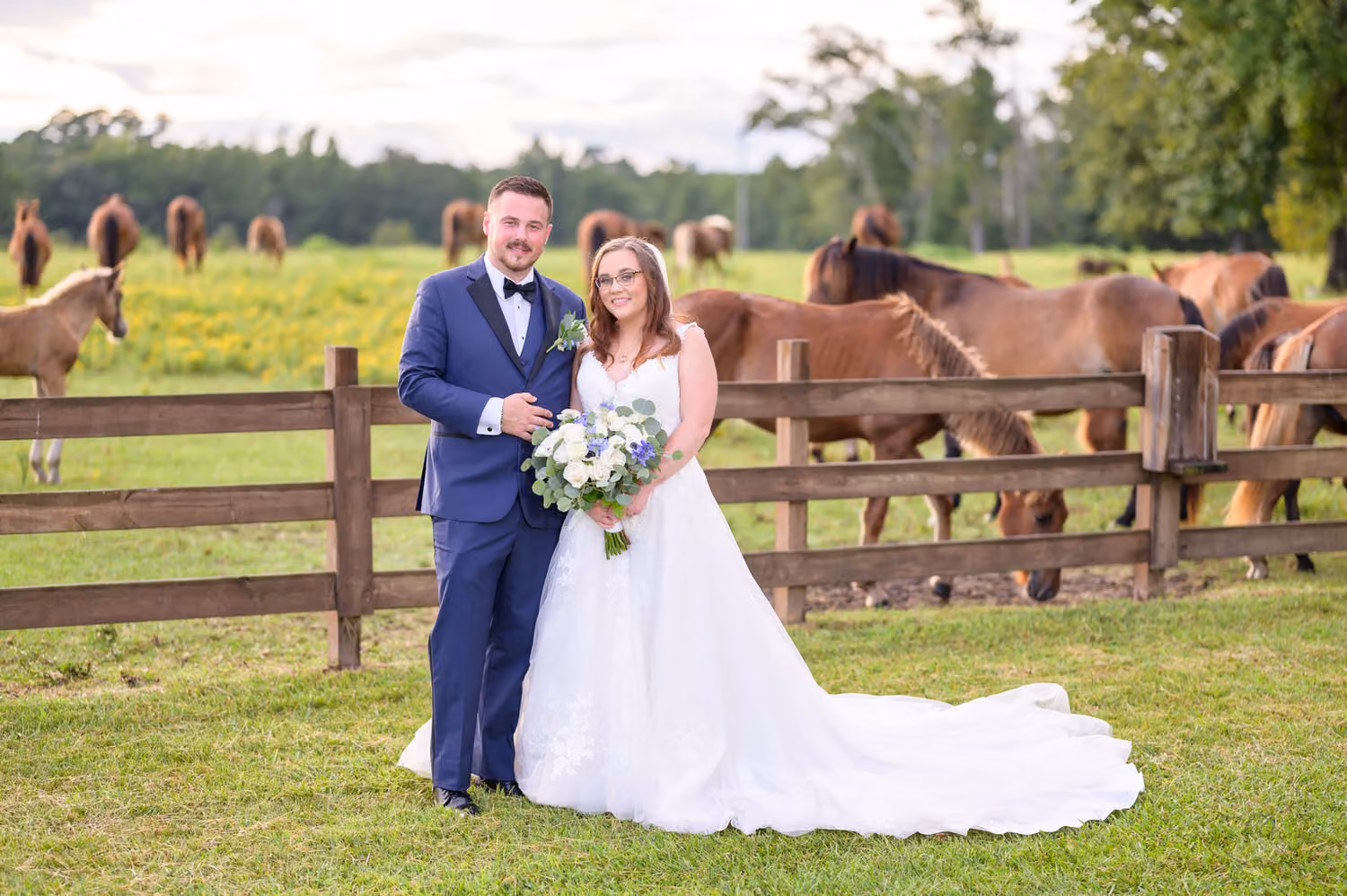Bride and groom in a blue suit standing by a wooden fence with horses grazing behind them at Wildhorse at Parker Farms