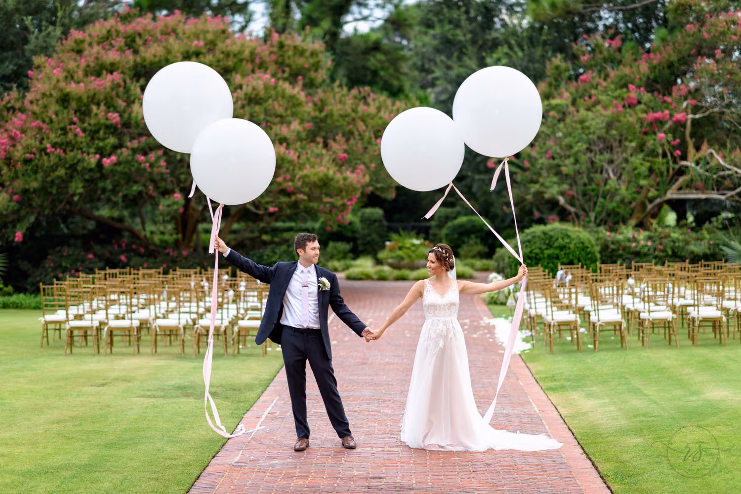 Bride and groom holding hands with big white balloons on a brick walkway by the ceremony chairs at Pine Lakes Country Club