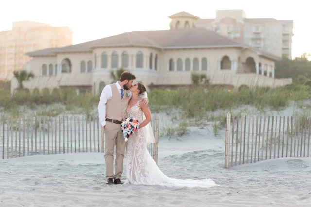 Bride and groom kissing on the beach with the Grande Dunes Ocean Club clubhouse behind them