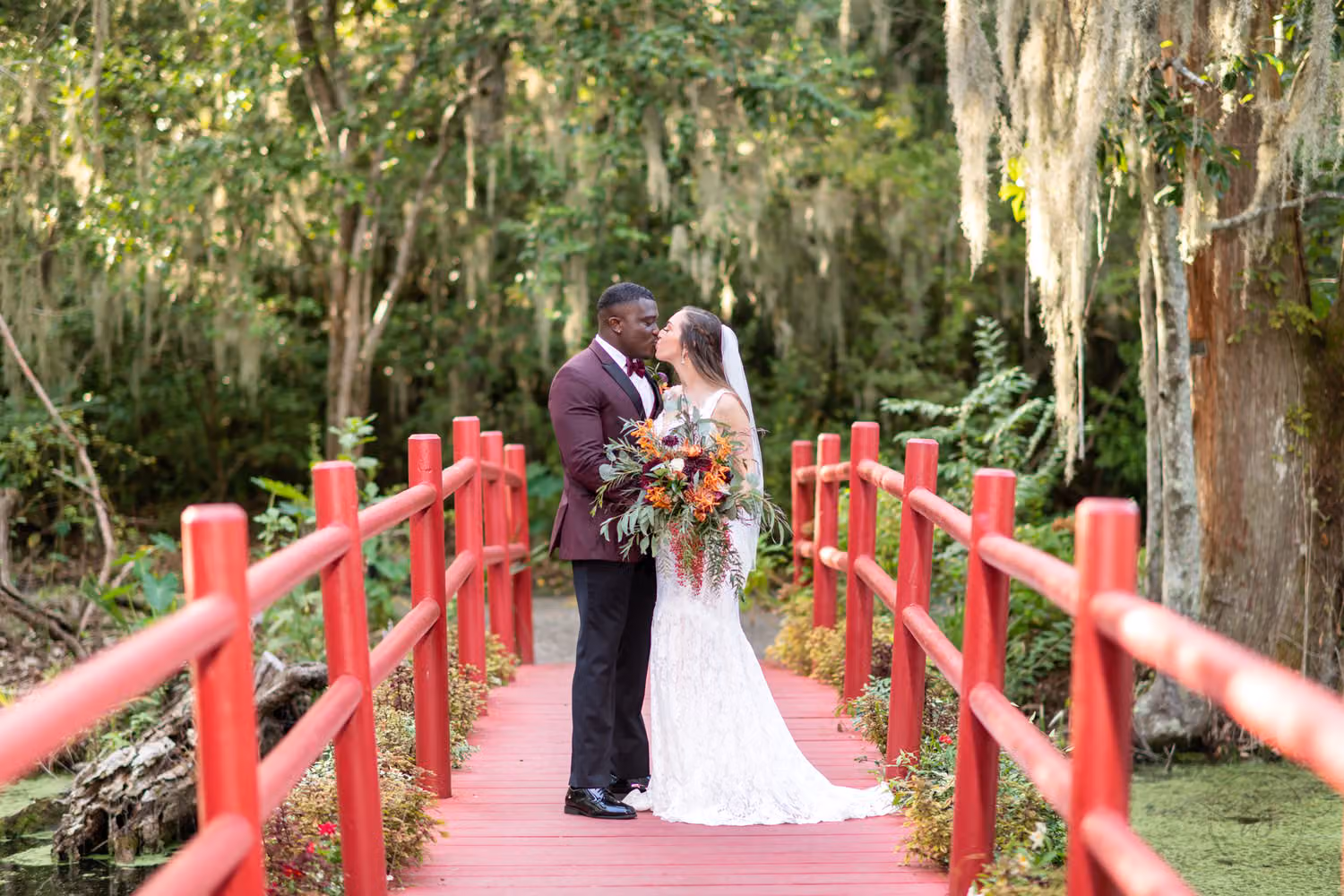Bride and groom kissing on a red wooden bridge surrounded by moss-draped trees at Magnolia Plantation in Charleston