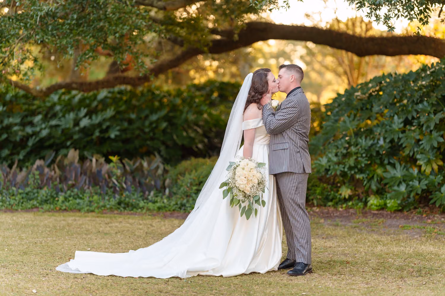 Bride with a long veil and groom in gray kissing on the lawn under spreading oak branches at Litchfield Country Club