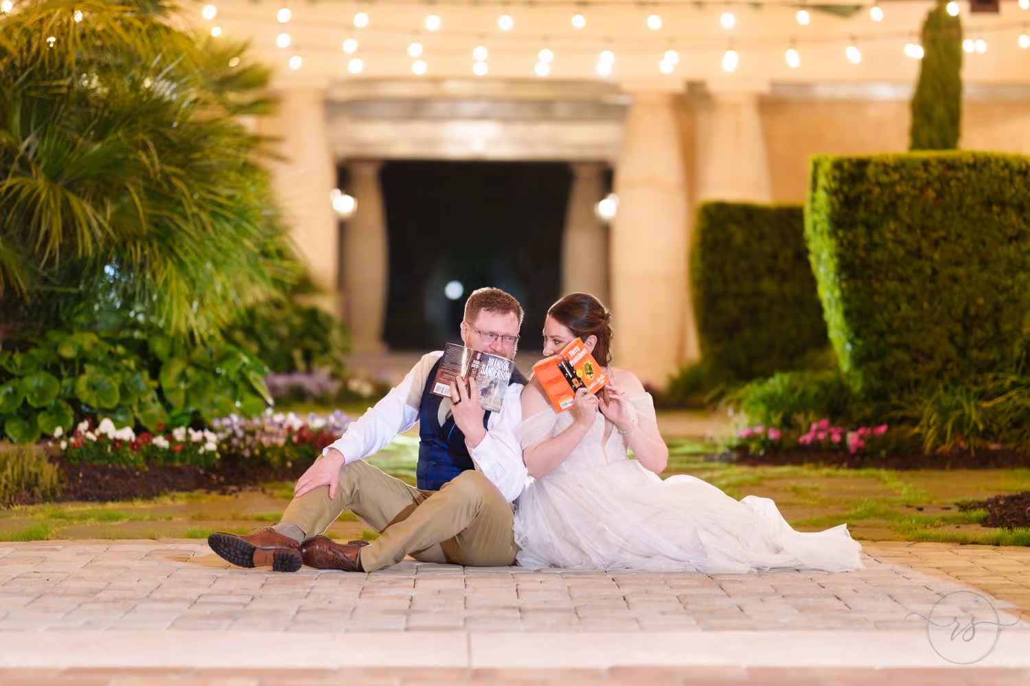 Bride and groom sitting on the courtyard pavement reading books under string lights at night at 21 Main Events