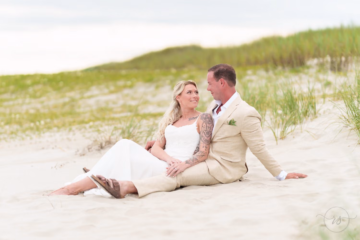 Bride and groom in a tan suit sitting together in the sand in front of grassy dunes on the beach