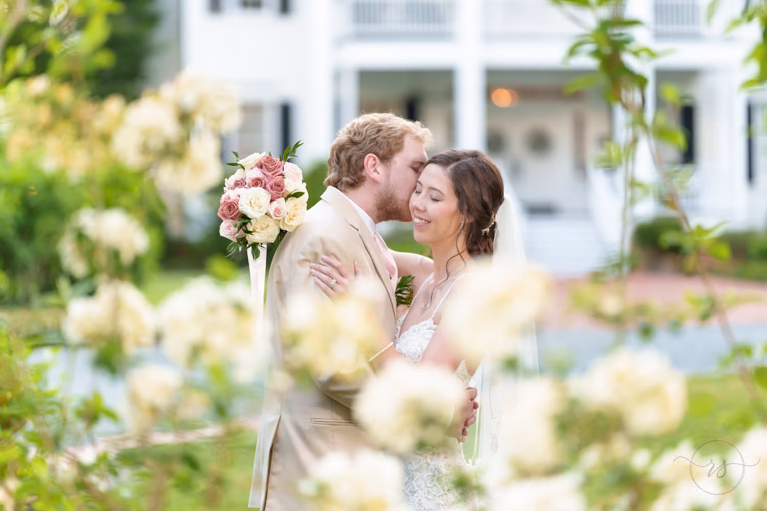 Bride and groom smiling close together behind white blooms in front of a white plantation house at Tanglewood Plantation