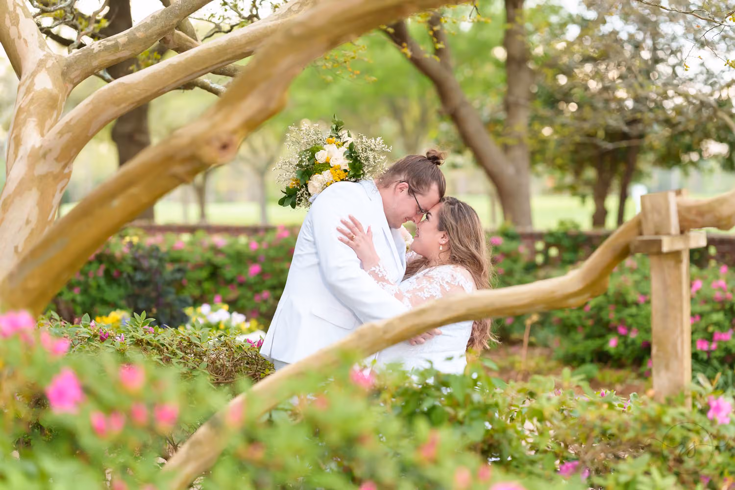Bride and groom in a white jacket touching foreheads behind crape myrtle branches and pink azaleas at Pine Lakes Country Club