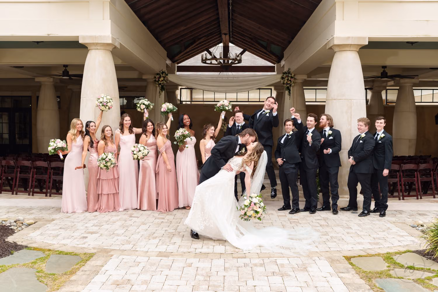 Groom dipping the bride as bridesmaids in pink and groomsmen in black cheer under the pavilion at 21 Main Events