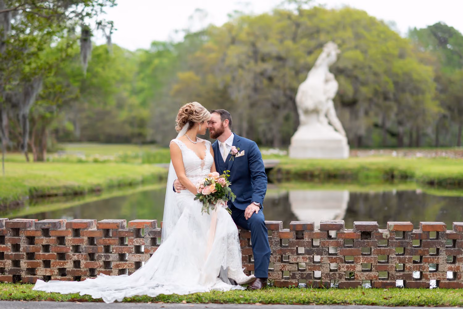 Bride and groom on a brick wall by the pond with the white Youth Taming the Wild sculpture behind them at Brookgreen Gardens