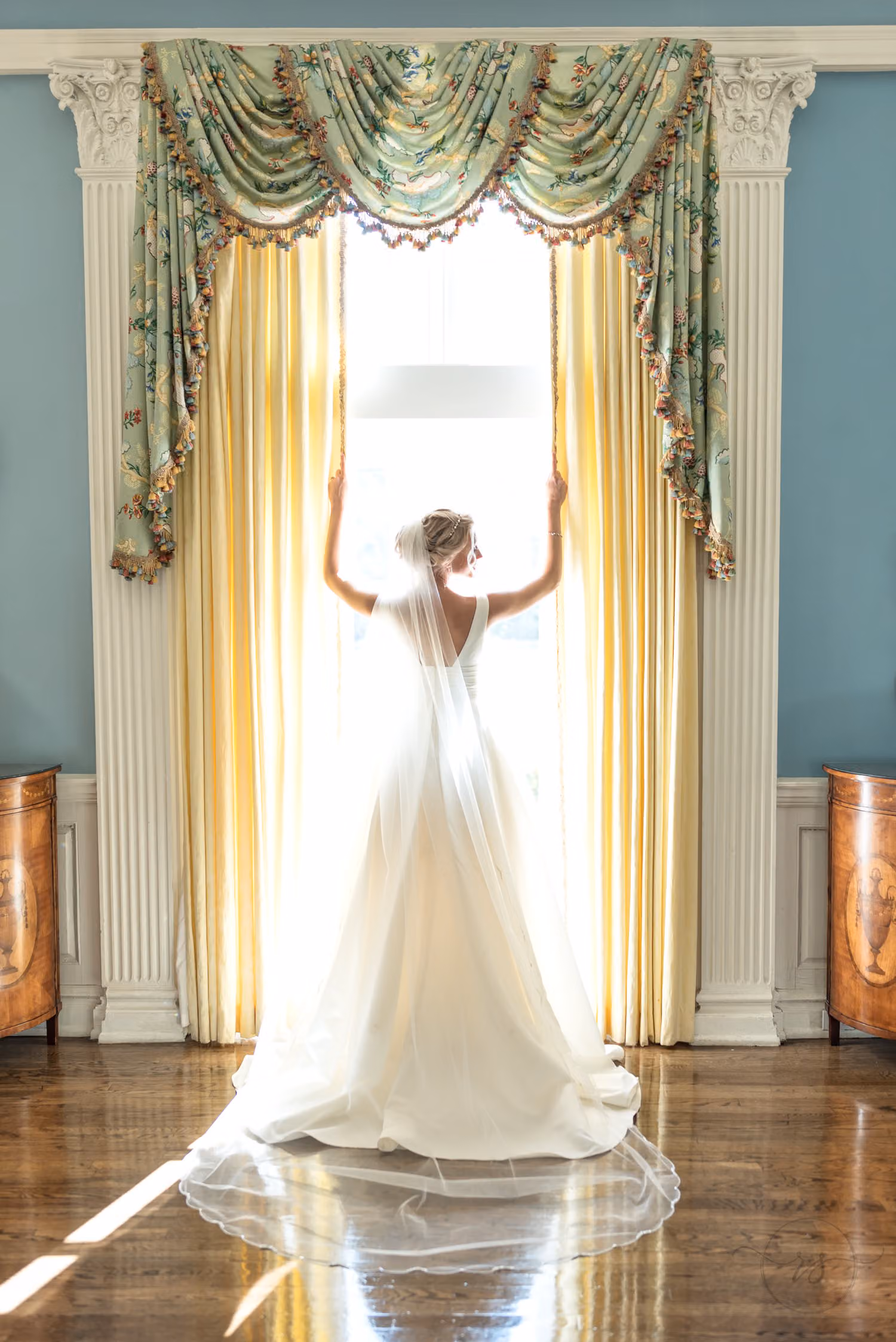 Bride seen from behind holding open tall curtains at a sunlit ballroom window at Pine Lakes Country Club