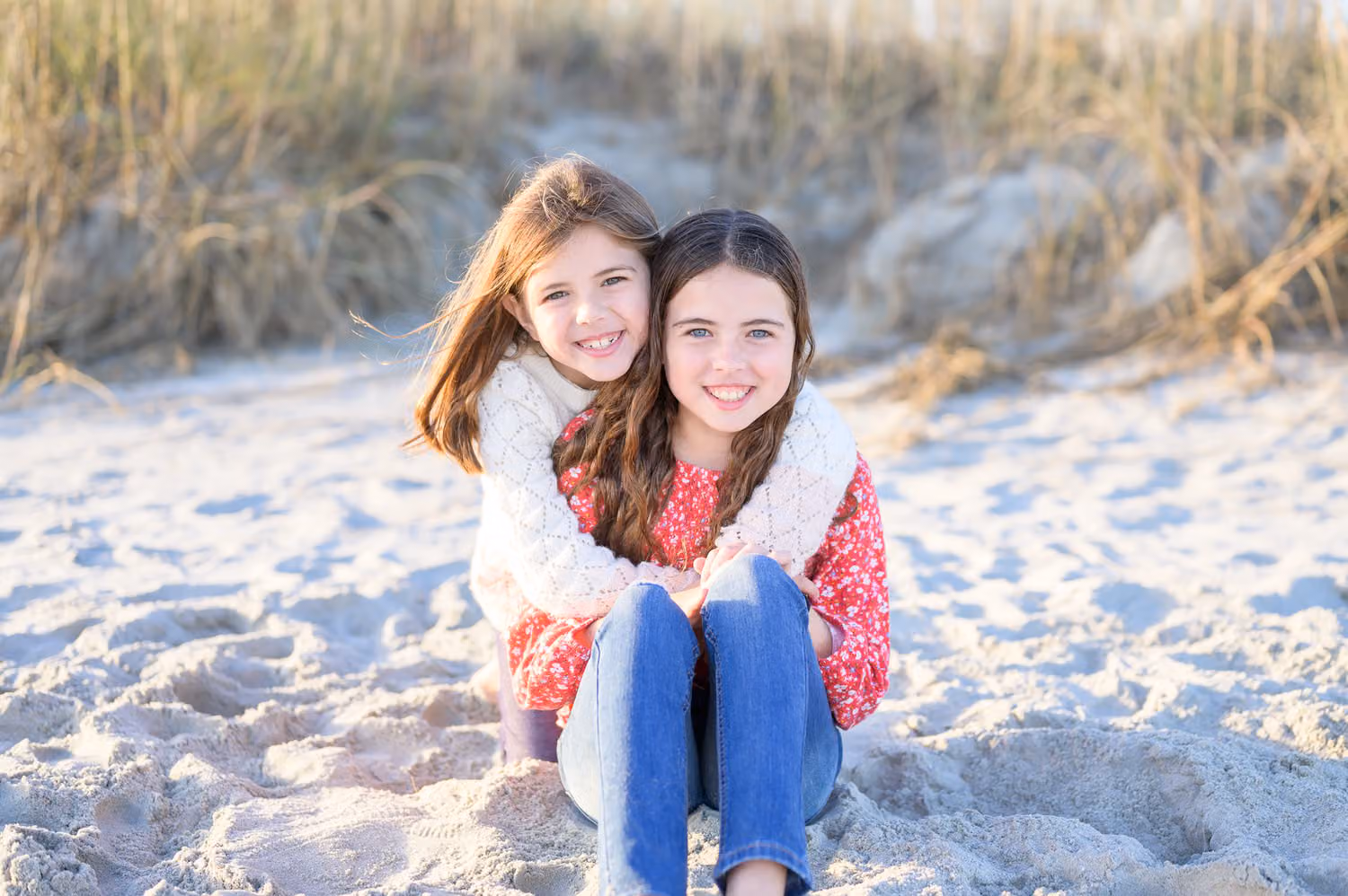 Two sisters hugging and smiling as they sit in the sand in front of the dune grass in North Myrtle Beach