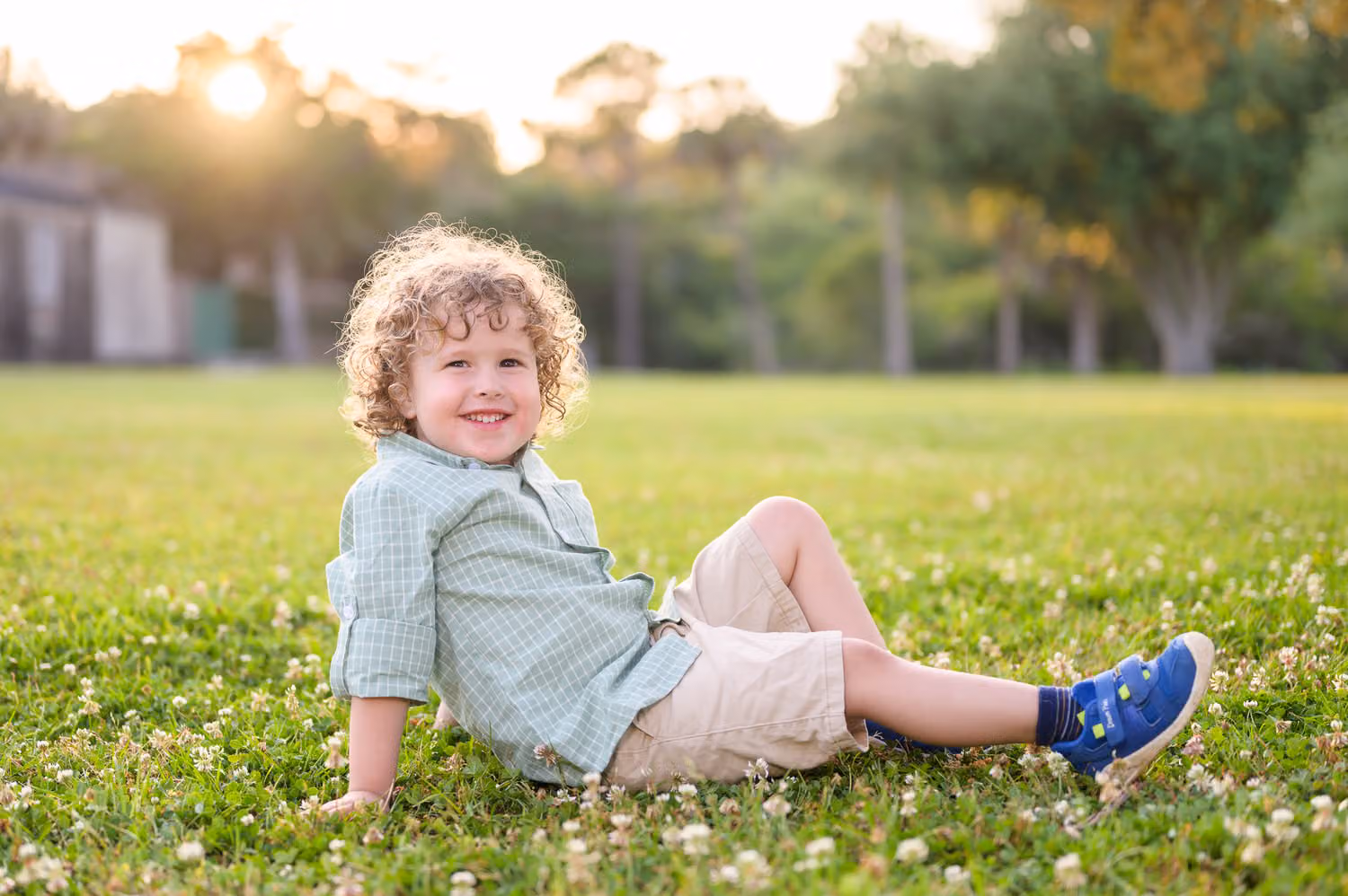 Curly-haired little boy lounging in the clover with the sun setting behind the trees at Huntington Beach State Park