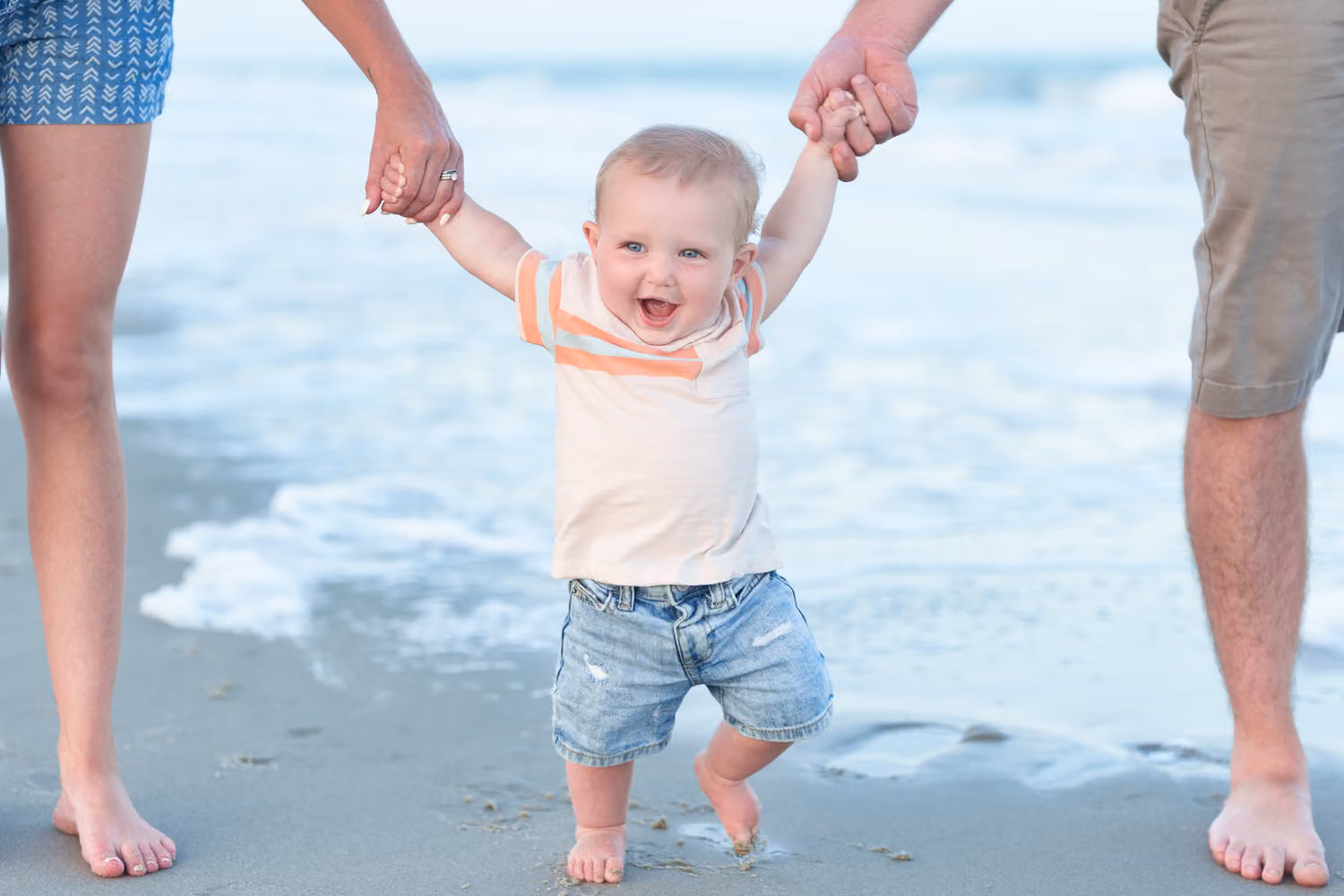 Laughing toddler boy taking steps at the water's edge while holding mom's and dad's hands at Huntington Beach State Park