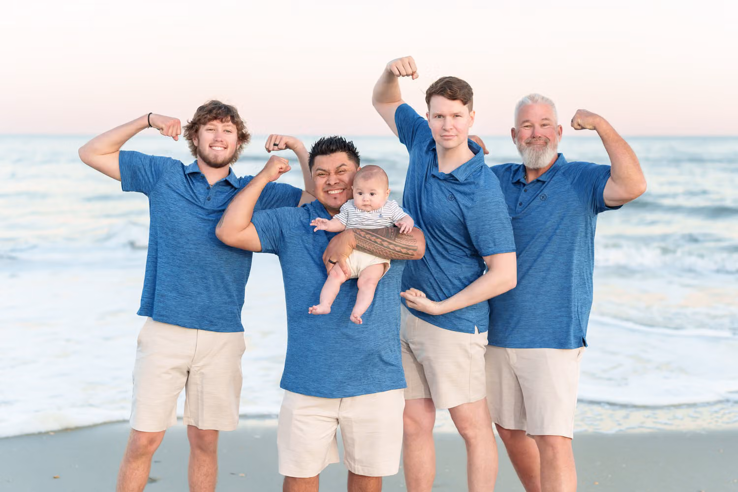 Four guys in matching blue shirts flexing their muscles, one holding a baby, on the beach at Huntington Beach State Park