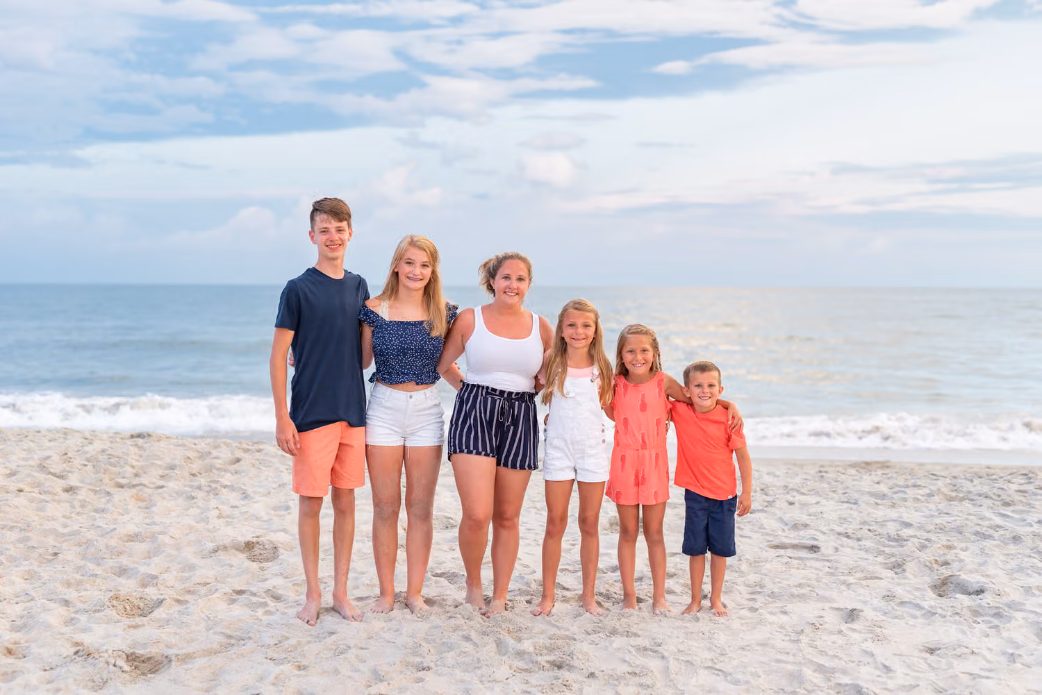 Six kids from teen to little girl lined up by height on the beach under blue sky and clouds at Huntington Beach State Park