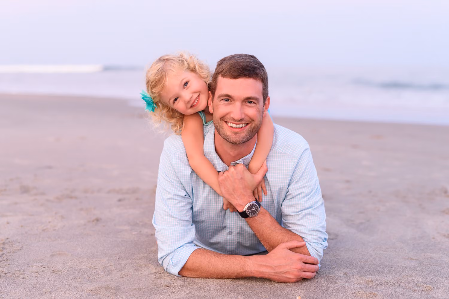 Little girl hugging her dad from behind as he lies smiling in the sand in Myrtle Beach