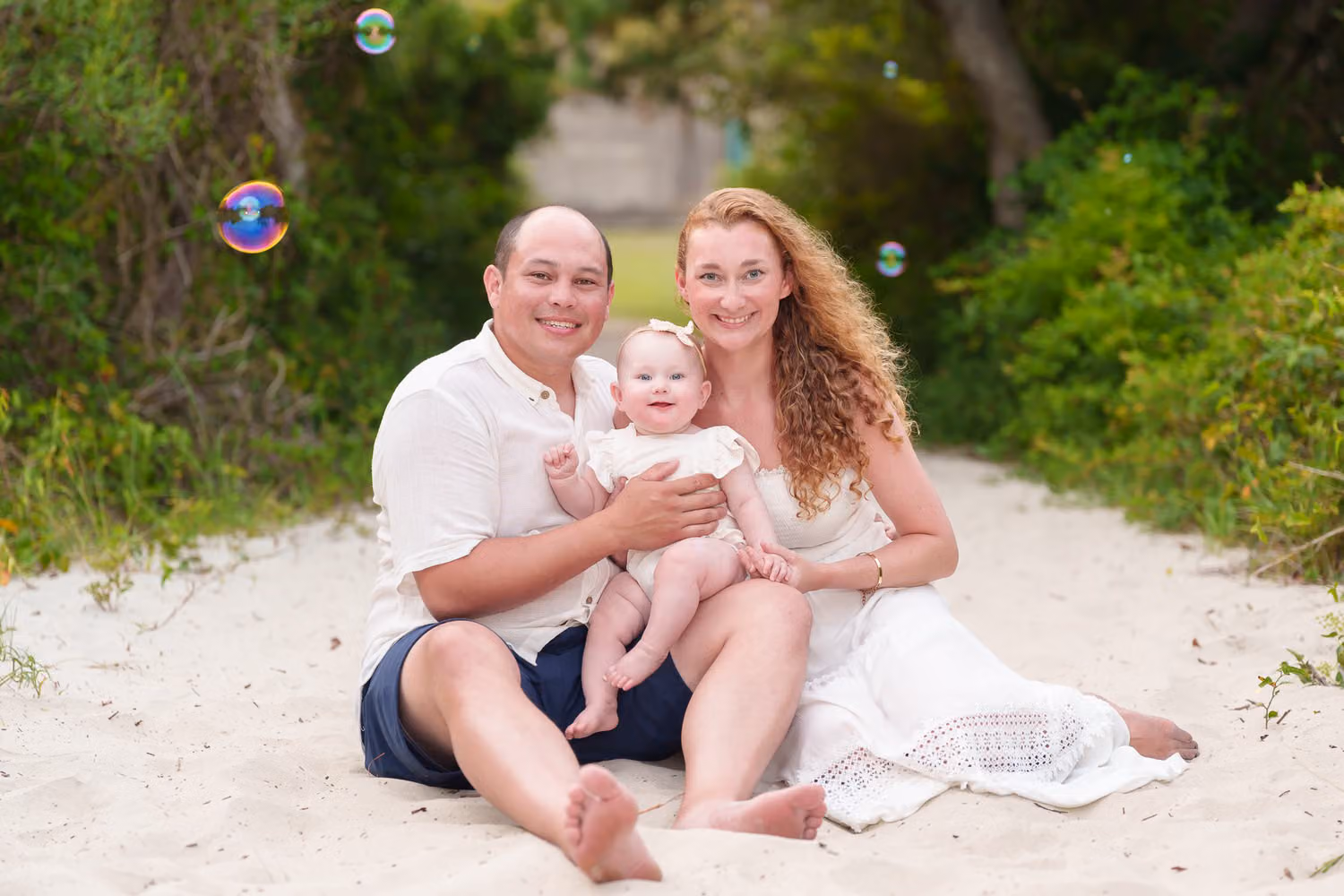 Mom and dad sitting with their baby on the sandy, tree-lined beach path as bubbles float by at Huntington Beach State Park