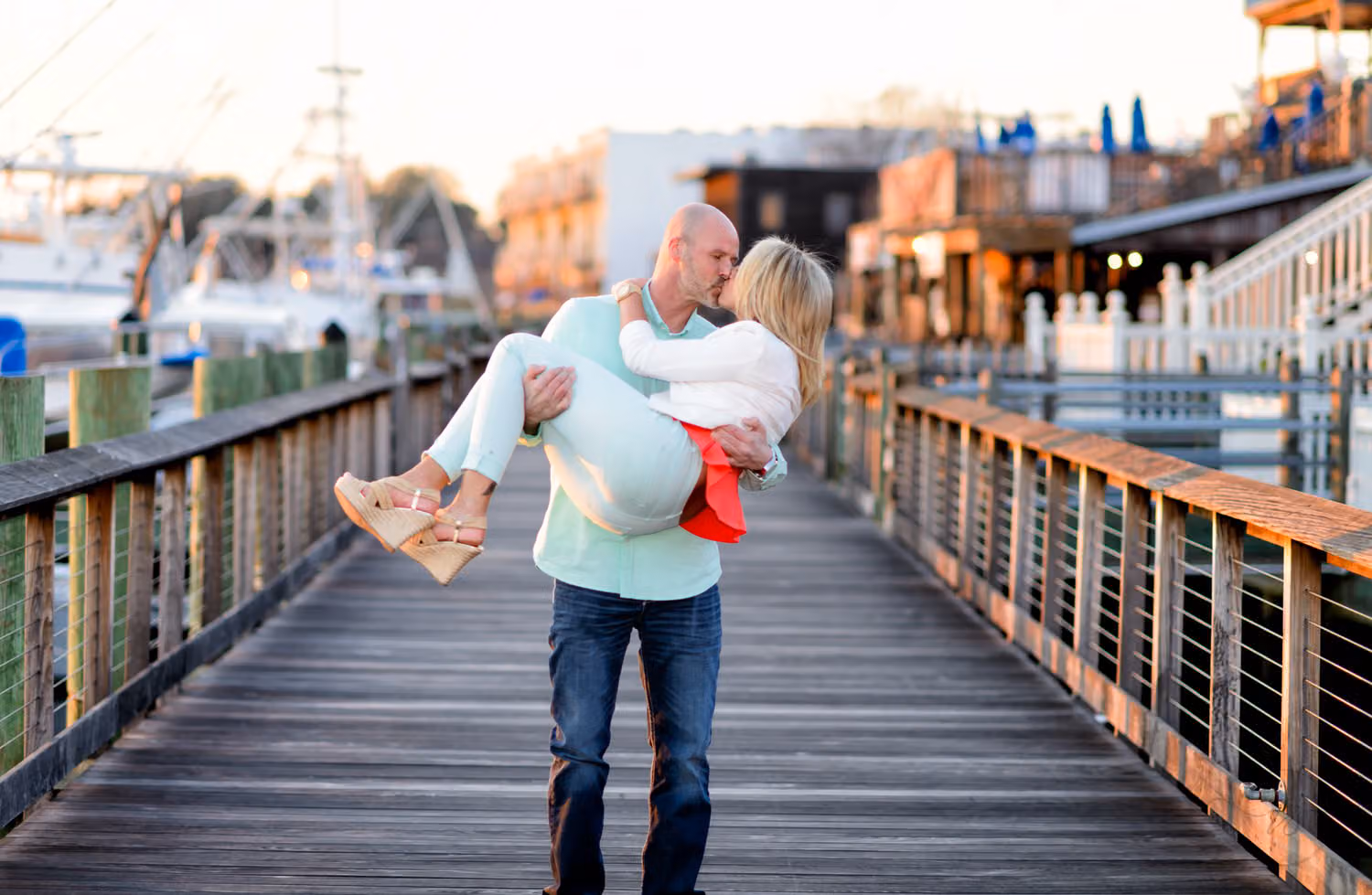 Man carrying his laughing fiancée in his arms down the wooden Harbor Walk past shops and boats in Downtown Georgetown